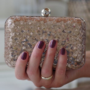 Könnte beinhalten: Eine goldene Clutch mit Pailletten und einem silbernen Verschluss. Die Clutch wird von einer Hand mit dunkelviolettem Nagellack gehalten.