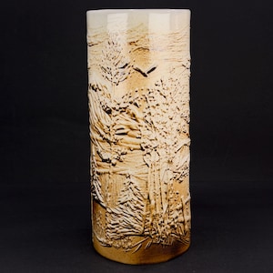 Puede incluir: Un jarrón cilíndrico de cerámica con un diseño de paisaje. El jarrón es de color marrón claro con una superficie texturizada que representa árboles y un pájaro en vuelo. El borde superior es de un tono más claro.