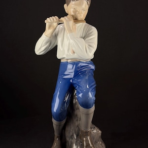 May include: A porcelain figurine of a boy playing a flute. The boy is wearing a white shirt, blue pants, and brown boots. The figurine is standing on a gray rock base. The boy's hair is brown.