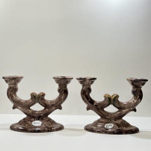 Puede incluir: Dos candelabros de cerámica marrón. Cada uno tiene dos copas para velas conectadas por un diseño curvo. Las bases son redondas y los candelabros tienen un acabado brillante. Accesorios decorativos para el hogar.