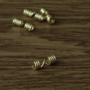 Könnte beinhalten: Mehrere kleine, goldfarbene Spiralperlen liegen auf einer Holzoberfläche. Die Perlen haben ein spiralförmiges Design, und die Holzmaserung bildet einen kontrastierenden Hintergrund. Sie könnten für die Schmuckherstellung oder andere Bastelarbeiten verwendet werden.