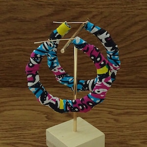 May include: A pair of large hoop earrings on a gold-coloured stand. The earrings are wrapped in fabric with a colourful pattern of pink, blue, black, and yellow. The stand is on a wooden surface.