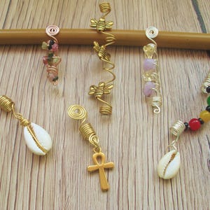 Puede incluir: Una colección de accesorios para el cabello hechos a mano, que incluyen envolturas de alambre dorado y plateado con cuentas, conchas y dijes. Los accesorios se exhiben en una clavija de madera sobre un fondo de vetas de madera clara.