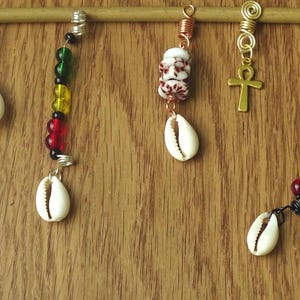 Può includere: Una collezione di accessori per capelli, tra cui conchiglie di ciprea, perline rosse, gialle, verdi e nere e un ciondolo Ankh. Gli accessori sono attaccati a un tassello di legno, mostrando vari design e materiali.
