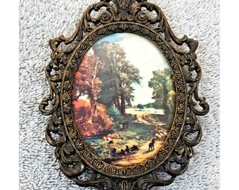 Puede incluir: Un marco de fotos ovalado con un diseño de metal dorado ornamentado. El marco contiene una impresión vintage de una escena de paisaje con árboles, un río y figuras.
