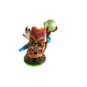 May include: A detailed figurine of a totem-like character with a red and brown face, green eyes, and a wide, toothy grin. It holds a green orb and has a feather and blue arm attached. The base is brown and green.
