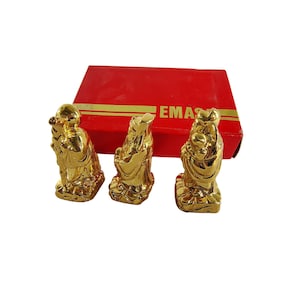 Puede incluir: Tres estatuas doradas de figuras en poses tradicionales, presentadas frente a una caja roja con letras doradas. Las estatuas son detalladas y parecen ser de metal. La caja tiene la palabra "EMAS" impresa.
