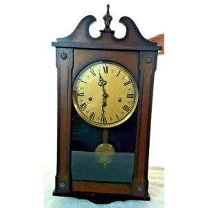 May include: A vintage wooden wall clock with a gold face and black Roman numeral hour markers. The clock has a pendulum and a glass cover.