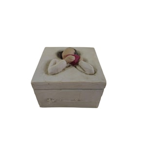 Könnte beinhalten: Eine cremefarbene dekorative Box mit einer geformten Figur, die ein rotes Herz hält. Die Figur sitzt, die Arme um das Herz gelegt. Die Box hat eine strukturierte Oberfläche und ein schlichtes, elegantes Design.