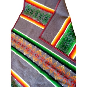 Puede incluir: Una manta tejida gris con un patrón geométrico colorido. El patrón presenta rayas de rojo, naranja, amarillo, verde y blanco. La manta tiene un ribete de cuero marrón.