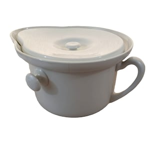 Puede incluir: Un orinal de cerámica blanca con tapa y asa. La tapa tiene un pequeño pomo en la parte superior.