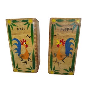May include: Wooden salt and pepper shakers with a rooster design. The shakers are rectangular and feature a painted rooster in orange, blue, and yellow. The word "Salt" is written on one shaker, and "Pepper" on the other.