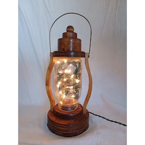 May include: A wooden lantern with a glass jar containing string lights. The lantern is made of dark wood and has a handle on top. The jar is clear glass and the lights are white. The lantern is sitting on a white surface.