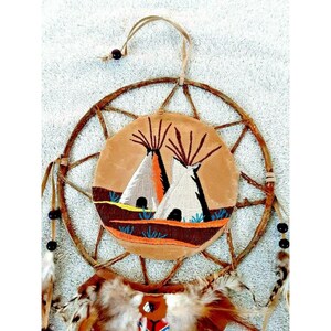 Puede incluir: Un atrapasueños de cuero marrón con flecos de cuentas y plumas. El atrapasueños tiene un marco de madera tejido y un círculo central con un diseño bordado de dos tipis.