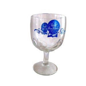 Puede incluir: Un vaso de cerveza de vidrio transparente con un logotipo azul de Pabst Blue Ribbon. El logotipo presenta una cinta azul con las palabras "Pabst Blue Ribbon Beer" impresas en él.