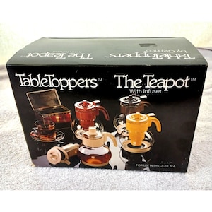 Könnte beinhalten: Eine Schachtel für eine Table Toppers The Teapot mit Infuser. Die Schachtel zeigt eine Auswahl an Teekannen in verschiedenen Farben, darunter Braun, Gelb und Weiß. Der Text auf der Schachtel lautet "Table Toppers The Teapot with Infuser" und "Zur Verwendung mit losem Tee."
