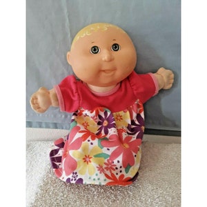 Puede incluir: Una muñeca Cabbage Patch Kid con cabello rubio y ojos verdes que lleva un vestido rosa con estampado floral.