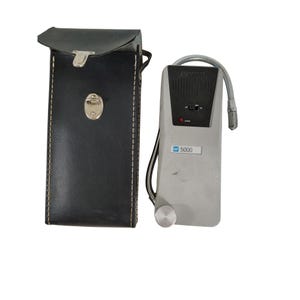 Puede incluir: Un dispositivo electrónico gris y negro vintage con un tubo metálico flexible y un estuche de transporte de cuero negro. El dispositivo tiene un dial, un altavoz y un indicador de encendido. El estuche es rectangular con un cierre plateado.