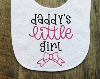 May include: White baby bib with the text "daddy's little girl" in black and pink lettering. A pink bow is below the text.
