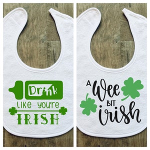 May include: Two white baby bibs with green shamrock designs and Irish-themed sayings. The first bib says "Drink like you're Irish" with a green baby bottle graphic. The second bib says "A wee bit Irish".