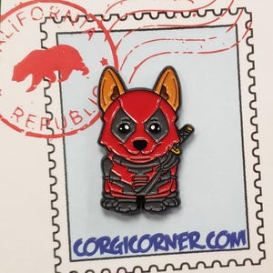 Op de afbeelding: Een rood en zwart geëmailleerde pin met een Corgi-hond in een Deadpool-kostuum, compleet met katana. De pin heeft een zwarte omtrek en staat tegen een lichtblauwe achtergrond met de tekst "CORGICORNER.COM".
