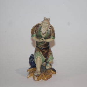 Op de afbeelding: Een keramieken beeldje van een oude man in traditionele kledij, zittend op een blauw en bruin vat. De figuur heeft een lange witte baard en draagt een groene en bruine jurk. De basis is een geel en bruin bladachtig figuur.