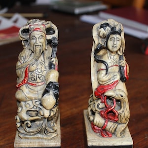 May include: Two ivory-coloured figurines of a man and a woman, both seated on a base. The man is wearing a red and gold robe and has a long white beard. The woman is wearing a red and white robe and has long black hair. Both figurines are decorated with intricate details.