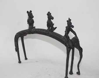 May include: A dark bronze sculpture depicting three figures riding atop a curved, arched structure. The figures appear to be riding animals. The sculpture has a matte finish and is set against a white background.