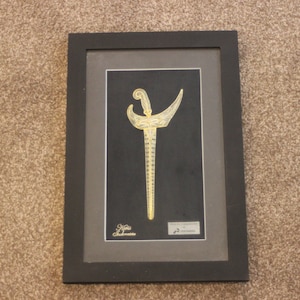 May include: A framed artwork featuring a gold-coloured dagger with intricate details against a black background. The frame is black with a grey inner border. The artwork is signed "Gift Souvenir" and includes the text "Kris" in a small white rectangle.