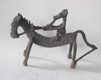 May include: A dark gray metal sculpture of a horse with a rider. The horse has a detailed mane and a patterned body. The rider is seated and holding the horse's reins. The sculpture is on a white background.