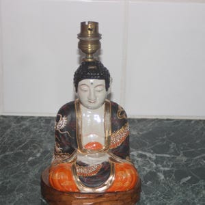 May include: A ceramic lamp base featuring a seated Buddha figure with intricate gold and black details. The Buddha is wearing a robe with orange and gold accents. The lamp base is made of wood and has a brass lampshade.