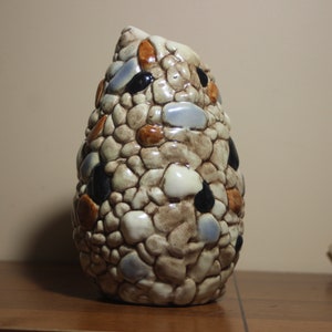 Peut inclure: Un vase en céramique avec une surface texturée unique, ressemblant à un groupe de petites pierres. Le vase est de couleur brun clair avec des accents de bleu, noir et orange.