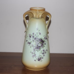 Puede incluir: Un jarrón de cerámica alto de color crema con un diseño floral. El jarrón tiene dos asas y un delicado patrón floral en tonos de morado y blanco.