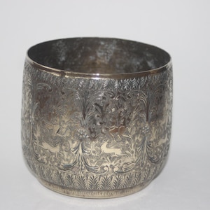 May include: A silver-coloured, cylindrical container with an intricate engraved design. The pattern features stylized animals and foliage. The vessel has a slightly flared rim and a rounded base. The overall aesthetic suggests a vintage or antique style.