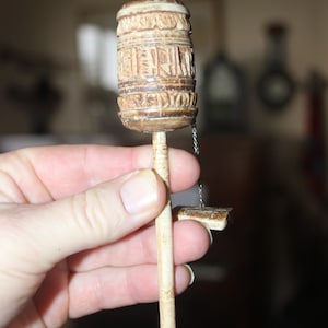 May include: A small, hand-held prayer wheel made of wood with intricate carvings. The wheel is attached to a long, thin wooden stick and has a small chain attached to the bottom.