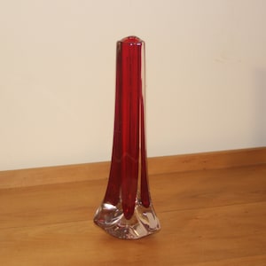 Könnte beinhalten: Eine hohe, schlanke, rote Glasvase mit einem ausgestellten Fuß. Die Vase ist aus klarem Glas mit einer roten Tönung.