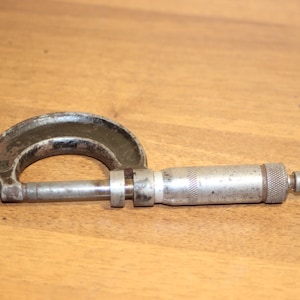 Könnte beinhalten: Ein silberner und schwarzer Mikrometer mit einem gerändelten Einstellknopf. Der Mikrometer wird verwendet, um die Dicke von Objekten zu messen.