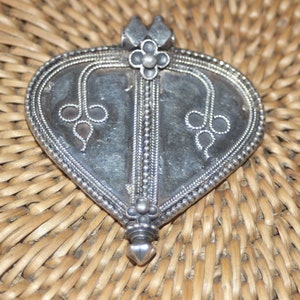 Peut inclure: Un pendentif en argent en forme de cœur avec des motifs floraux complexes et une bordure perlée. Le pendentif a une pointe pointue et une boucle en haut pour attacher une chaîne.