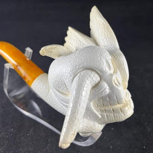 Puede incluir: Una pipa de tabaco tallada con un diseño de calavera, alas y serpiente. La pipa tiene una boquilla marrón claro y un cuenco blanco. La pipa se exhibe sobre un soporte transparente sobre un fondo oscuro.