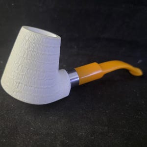 Peut inclure: Une pipe à tabac blanche avec un foyer texturé et un tuyau incurvé orange. La pipe a une bague argentée reliant le foyer et le tuyau. La pipe est posée sur un fond sombre.