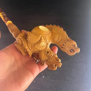 May include: A carved wooden pipe featuring a detailed dragon design. The pipe has a light brown color with intricate carvings of dragon heads and scales. The stem is a darker brown with yellow accents.