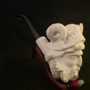 May include: A white meerschaum tobacco pipe featuring a detailed carving of a bearded figure wearing a horned helmet. The pipe has a smooth, black acrylic stem and is sculpted with intricate textures on the helmet and the facial hair of the figure.