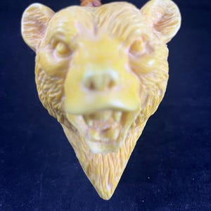 May include: A detailed, yellow-toned bear head carving with visible teeth and textured fur. The bear's head is carved with a triangular base and a small, brown, cylindrical piece at the top. The carving is set against a dark background.