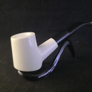 May include: A white ceramic pipe with a black stem and a curved design. The pipe is resting on a black stand, set against a dark background. The pipe's bowl is cylindrical, and the stem is curved.