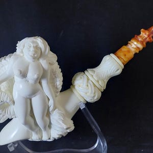 May include: A cream-colored tobacco pipe featuring a carved nude female figure. The pipe has a curved stem with a brown and tan mouthpiece. The bowl is adorned with intricate carvings. The pipe is displayed on a clear stand against a dark background.