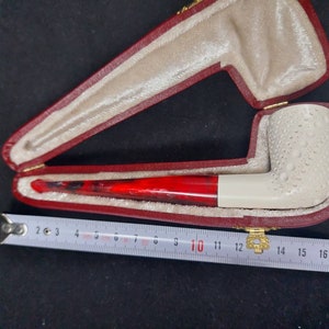 May include: A white tobacco pipe with a red and black stem, resting in an open red case with a cream-colored interior. The pipe has a textured surface. A measuring tape is next to the case.