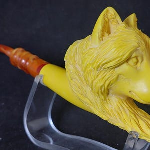 May include: A yellow tobacco pipe with a carved wolf head design. The pipe has a yellow bowl, a brown and orange stem, and a curved mouthpiece. The wolf head is detailed with fur and facial features.