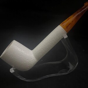 Puede incluir: Una pipa de tabaco blanca con un patrón floral texturizado en el cuenco y el vástago. La boquilla es de color marrón veteado. La pipa se exhibe sobre un soporte transparente sobre un fondo negro.