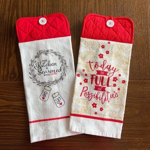 May include: Two white kitchen towels with red quilted pot holders. The first towel has a wreath with the text "This Kitchen is Seasoned with Love" and two salt and pepper shakers. The second towel has the text "Today is Full of Possibilities" with red flowers.