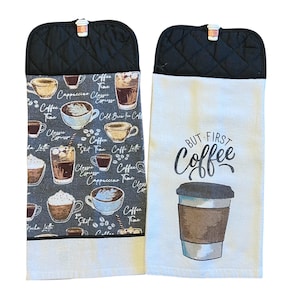 May include: Two hanging kitchen towels. One features coffee cup illustrations and text, the other says "BUT FIRST COFFEE" with a coffee cup graphic. Both have black quilted tops and a clip.